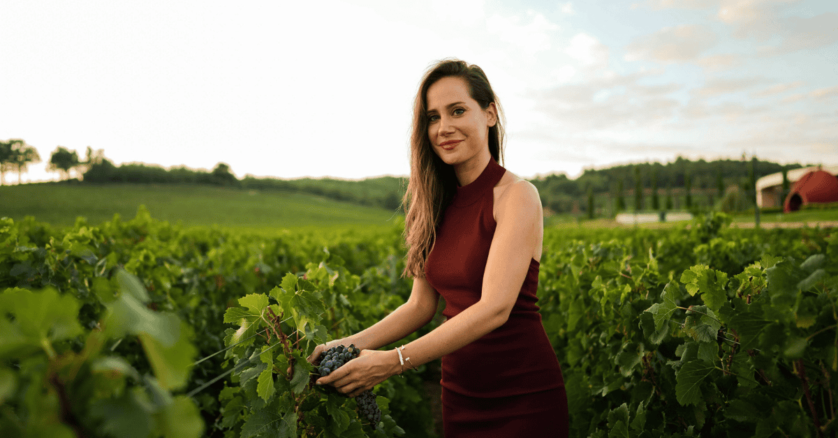 Woman in elegant midi dress standing in vineyard holding grapes during golden hour wine tasting